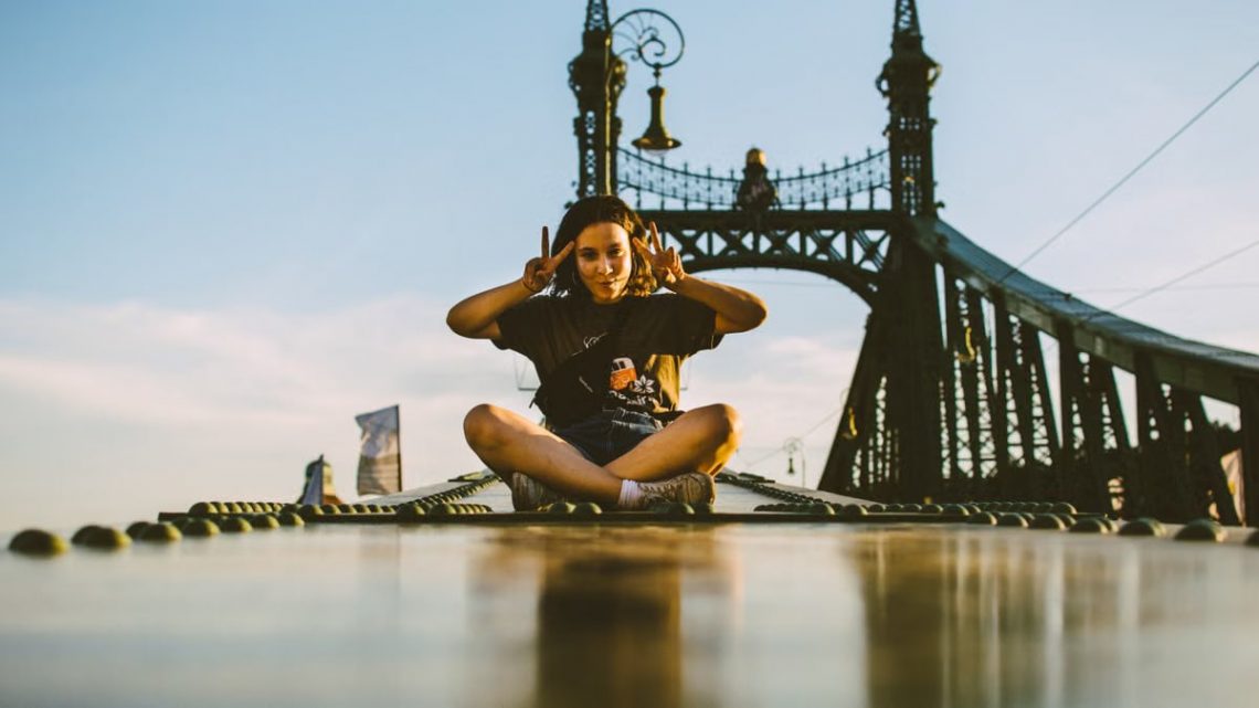 A young tourist poses with a peace sign while seated cross-legged on Budapest's Liberty Bridge during a sunny day.