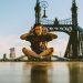 A young tourist poses with a peace sign while seated cross-legged on Budapest's Liberty Bridge during a sunny day.